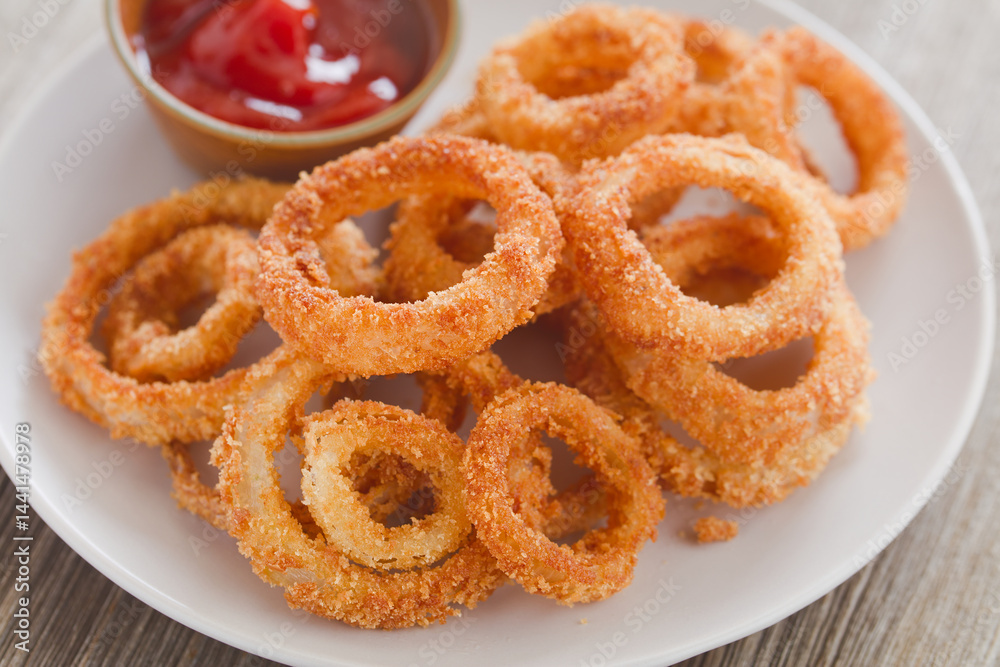 Homemade crispy fried breaded onion rings on a plate with a bowl of ketchup (Selective Focus, Focus on parts of the onion rings in the front) Homemade crispy fried breaded onion rings on a plate with a bowl of ketchup (Selective Focus, Focus on parts of the onion rings in the front)