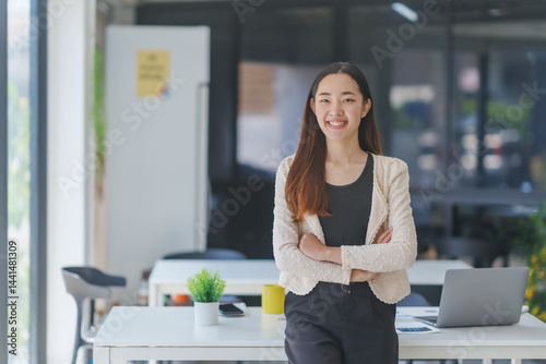 Portrait of a smiling asian businesswoman standing with arms crossed in a modern office, showing confidence and professionalism