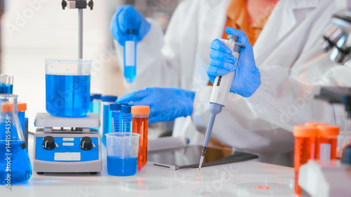 Obraz na płótnie Two Asian female scientists wearing laboratory gloves sit at a table, carefully examining a test tube filled with blue liquid, using a microscope for detailed analysis in a research laboratory w salonie