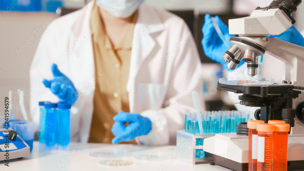 Two Asian female scientists wearing laboratory gloves sit at a table, carefully examining a test tube filled with blue liquid, using a microscope for detailed analysis in a research laboratory
