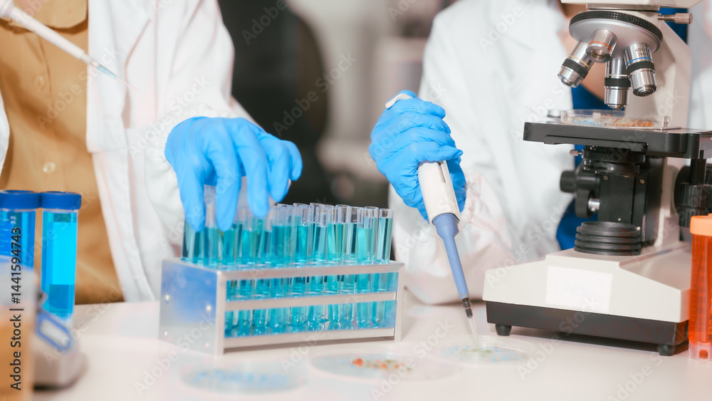Two Asian female scientists wearing laboratory gloves sit at a table, carefully examining a test tube filled with blue liquid, using a microscope for detailed analysis in a research laboratory