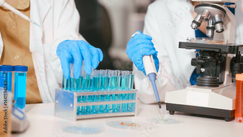 Obraz na płótnie Two Asian female scientists wearing laboratory gloves sit at a table, carefully examining a test tube filled with blue liquid, using a microscope for detailed analysis in a research laboratory w salonie