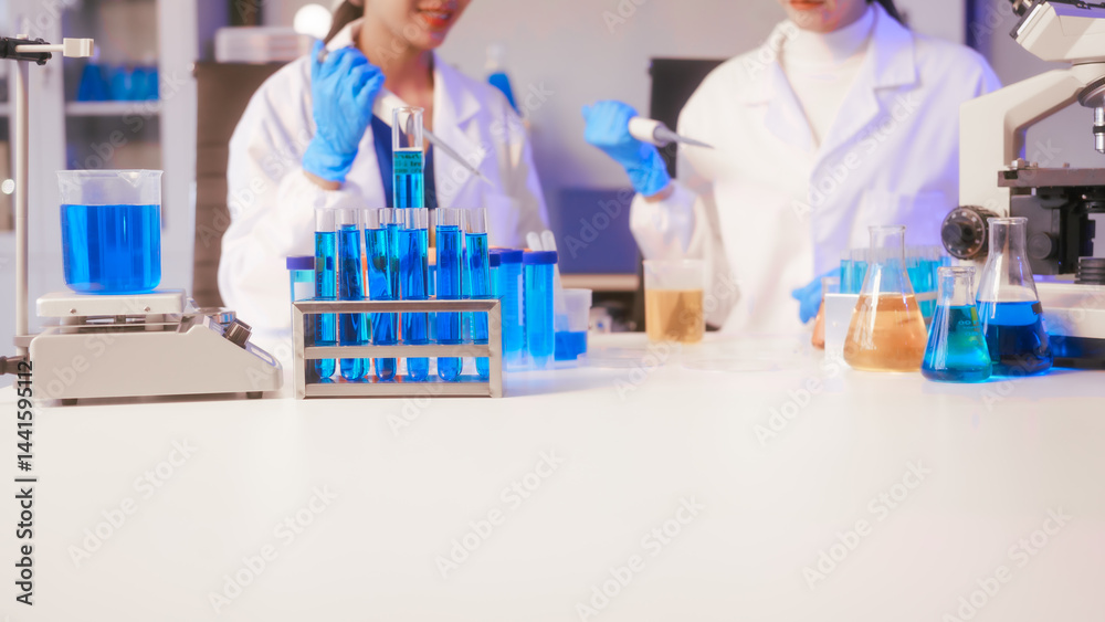 Two Asian female scientists wearing laboratory gloves sit at a table, carefully examining a test tube filled with blue liquid, using a microscope for detailed analysis in a research laboratory
