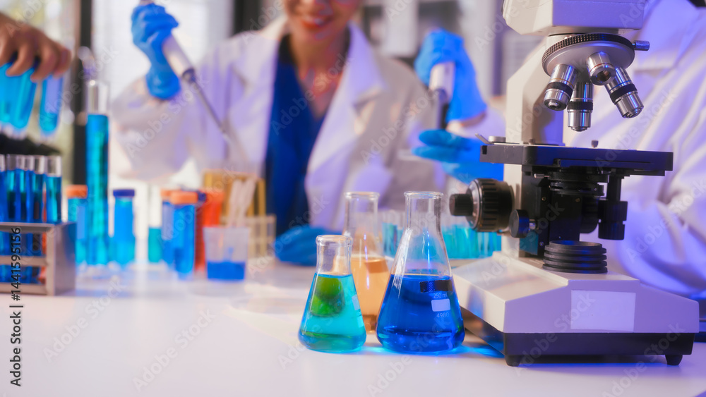 Two Asian female scientists wearing laboratory gloves sit at a table, carefully examining a test tube filled with blue liquid, using a microscope for detailed analysis in a research laboratory