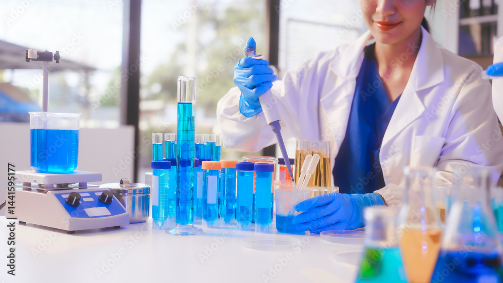 Fototapeta premium Two Asian female scientists wearing laboratory gloves sit at a table, carefully examining a test tube filled with blue liquid, using a microscope for detailed analysis in a research laboratory