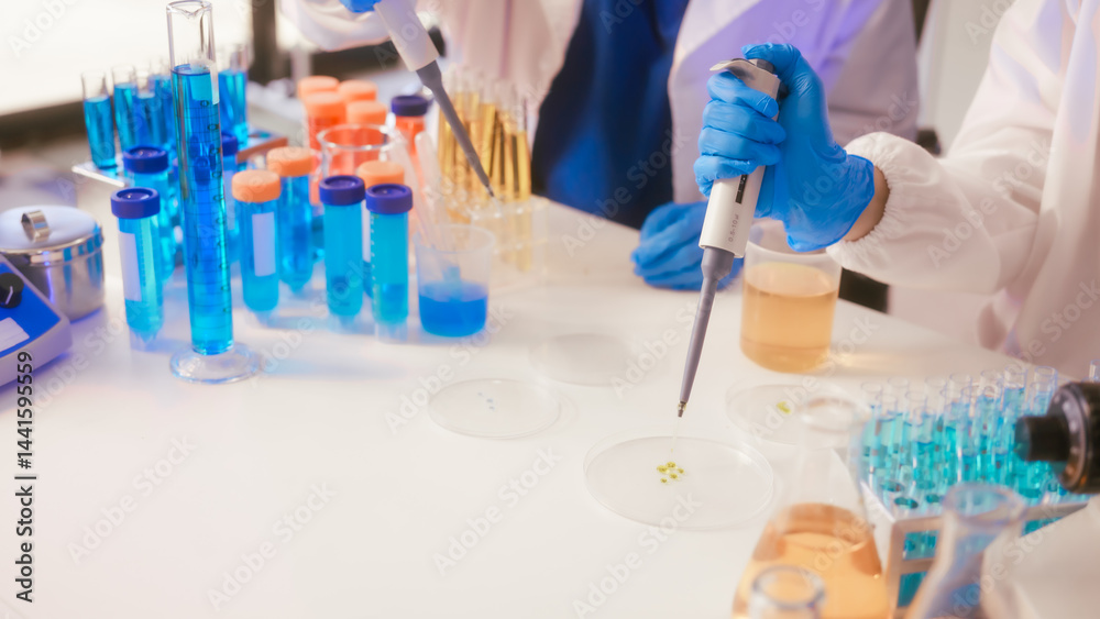 Two Asian female scientists wearing laboratory gloves sit at a table, carefully examining a test tube filled with blue liquid, using a microscope for detailed analysis in a research laboratory