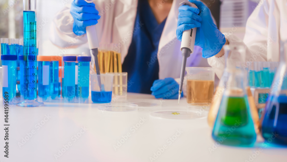 Two Asian female scientists wearing laboratory gloves sit at a table, carefully examining a test tube filled with blue liquid, using a microscope for detailed analysis in a research laboratory