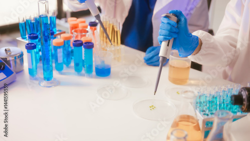 Obraz na płótnie Two Asian female scientists wearing laboratory gloves sit at a table, carefully examining a test tube filled with blue liquid, using a microscope for detailed analysis in a research laboratory w salonie