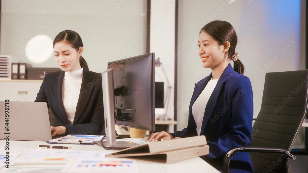 Two young Asian business woman sit at a table in the office, discussing marketing strategies, using a computer reviewing documents with charts and graphs, collaborating on new business plans at work