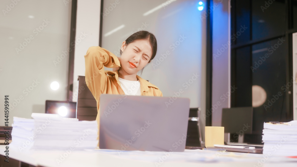 An Asian woman works at her office desk, feeling stressed and experiencing pain from sitting for a long time, struggling with discomfort and fatigue while trying to stay focused on her tasks.