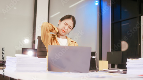 Obraz na płótnie An Asian woman works at her office desk, feeling stressed and experiencing pain from sitting for a long time, struggling with discomfort and fatigue while trying to stay focused on her tasks. w salonie