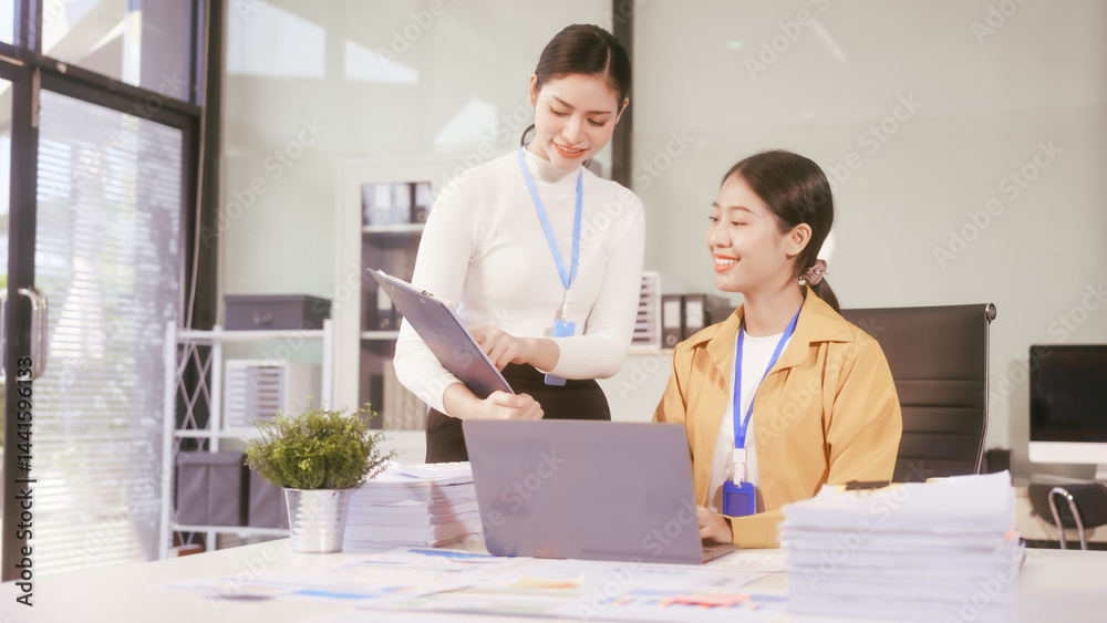 Two young Asian business woman sit at a table in the office, discussing marketing strategies, using a computer reviewing documents with charts and graphs, collaborating on new business plans at work