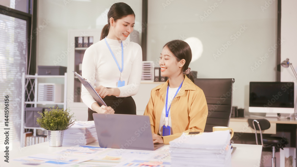 Two young Asian business woman sit at a table in the office, discussing marketing strategies, using a computer reviewing documents with charts and graphs, collaborating on new business plans at work
