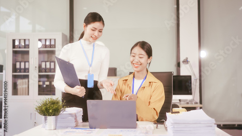 Two young Asian business woman sit at a table in the office, discussing marketing strategies, using a computer reviewing documents with charts and graphs, collaborating on new business plans at work
