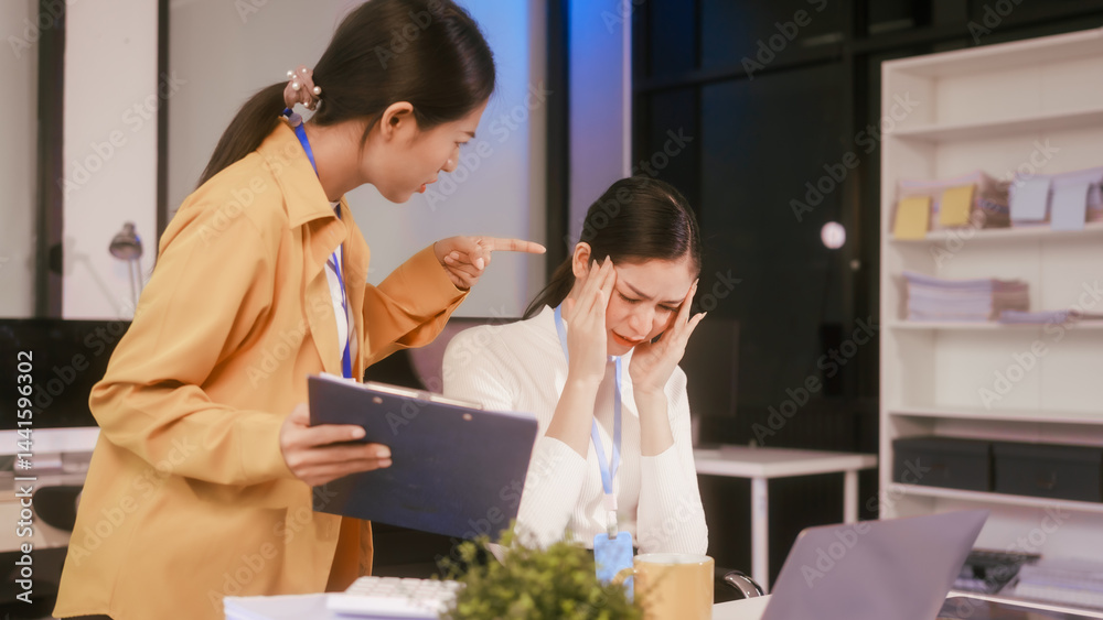 Two Asian women sit working at desk,overwhelmed with stress struggling with headaches,feeling mentally and physically exhausted while trying to concentrate on their demanding tasks in a busy office