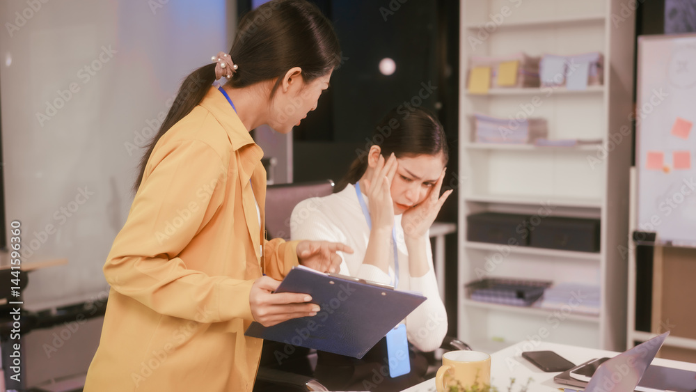 Two Asian women sit working at desk,overwhelmed with stress struggling with headaches,feeling mentally and physically exhausted while trying to concentrate on their demanding tasks in a busy office