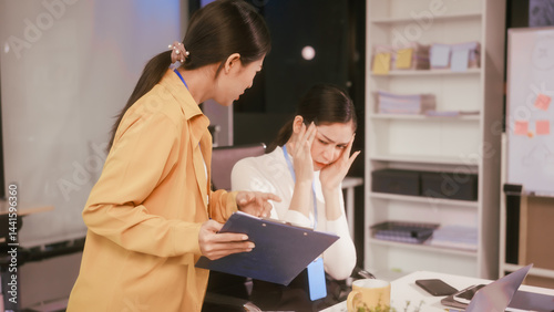 Obraz na płótnie Two Asian women sit working at desk,overwhelmed with stress struggling with headaches,feeling mentally and physically exhausted while trying to concentrate on their demanding tasks in a busy office w salonie