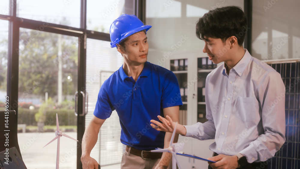 Two Asian businessmen wearing engineer hats table, discussing solar panels and wind turbines. They plan environmental projects, focusing on solar energy solutions and renewable energy installations
