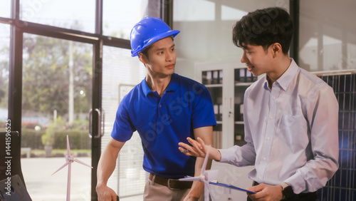 Obraz na płótnie Two Asian businessmen wearing engineer hats table, discussing solar panels and wind turbines. They plan environmental projects, focusing on solar energy solutions and renewable energy installations w salonie