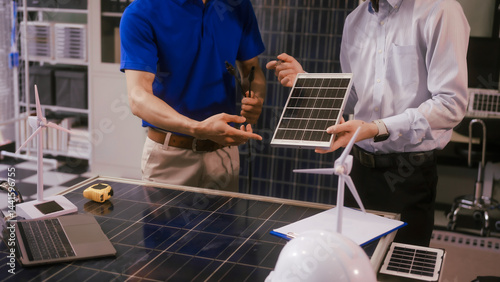 Obraz na płótnie Two Asian businessmen wearing engineer hats table, discussing solar panels and wind turbines. They plan environmental projects, focusing on solar energy solutions and renewable energy installations w salonie