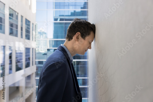Фотография Young 30s businessman leaning against wall his head, feeling exhausted and overwhelmed, experiences emotional challenges faced in corporate world