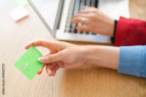 A person is shopping online using a laptop. They are holding a green credit card, likely to make a purchase.