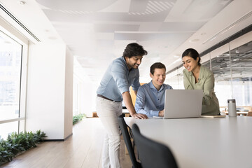 Three multinational colleagues gathered around laptop in modern coworking space, collaborate on project, discuss ideas, assign tasks and work on shared goal during briefing event in corporate office