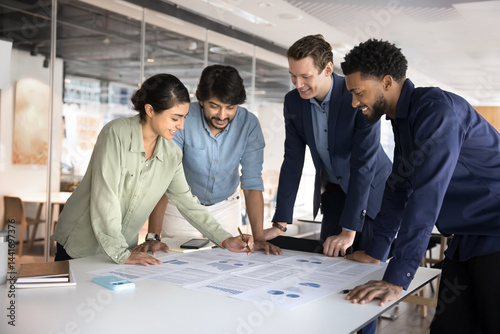 Group of smiling multiethnic people reviewing documents, analyzing infographic data, engaged in collaborative discussion, learning charts and graphs together, gathered around table in modern office