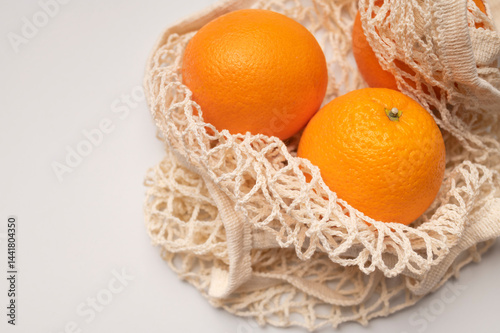 Ripe juicy oranges in cotton string bag on light gray background. Eco-friendly thread bag for shopping, buying food, fruits, vegetables. Zero waste, plastic free concept 