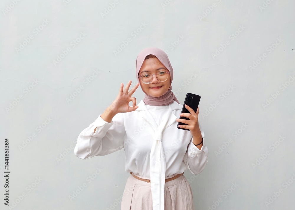 Fototapeta premium Smiling Young Muslim Woman in Hijab Showing OK Hand Gesture While Holding Smartphone Against Plain Background