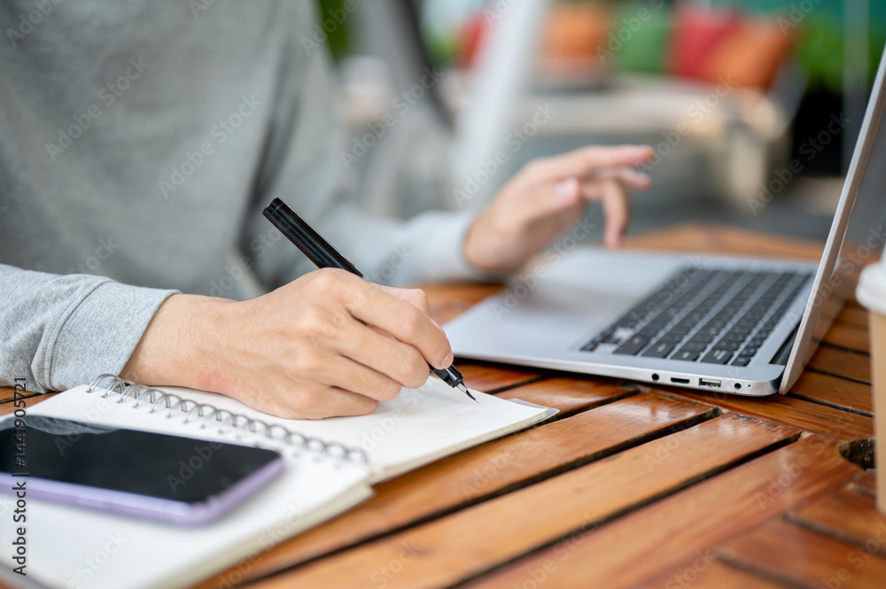 A hand holding a pen writing in notebook and other hand hovering over laptop sitting at wooden table