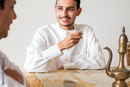 Two men in traditional attire enjoy coffee. One man holds a cup, smiling. Traditional coffee pot on table. Cultural coffee setting with traditional elements. Muslim young man drinking tea. photo