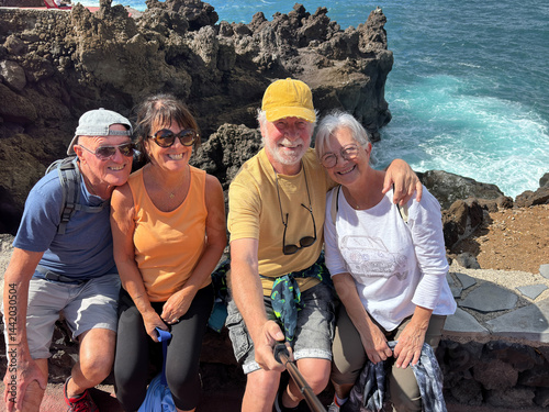 Four cheerful seniors along the sea coast looking at phone for selfie - Group of smiling pensioners enjoying nature and taking a walk together on a sunny day
