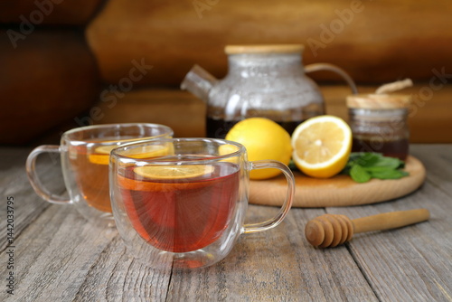 On an old wooden table against a log wall, in front of a tray with a glass teapot, lemon, mint and a glass jar of honey, there are two glass cups of hot black tea.