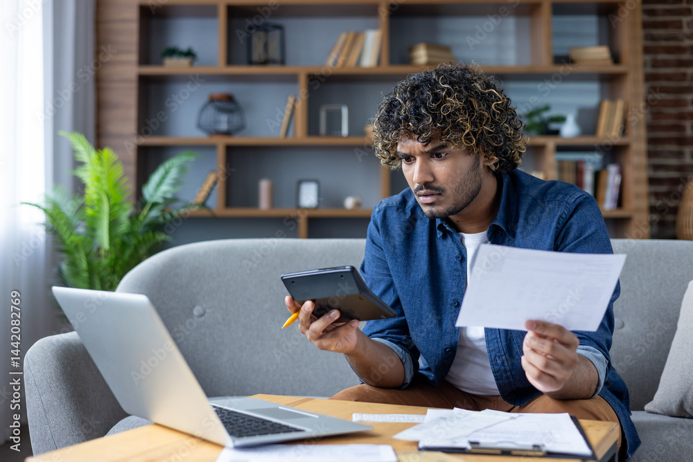 A concerned man calculates finances with a calculator and paperwork while using a laptop at home. The man appears stressed.