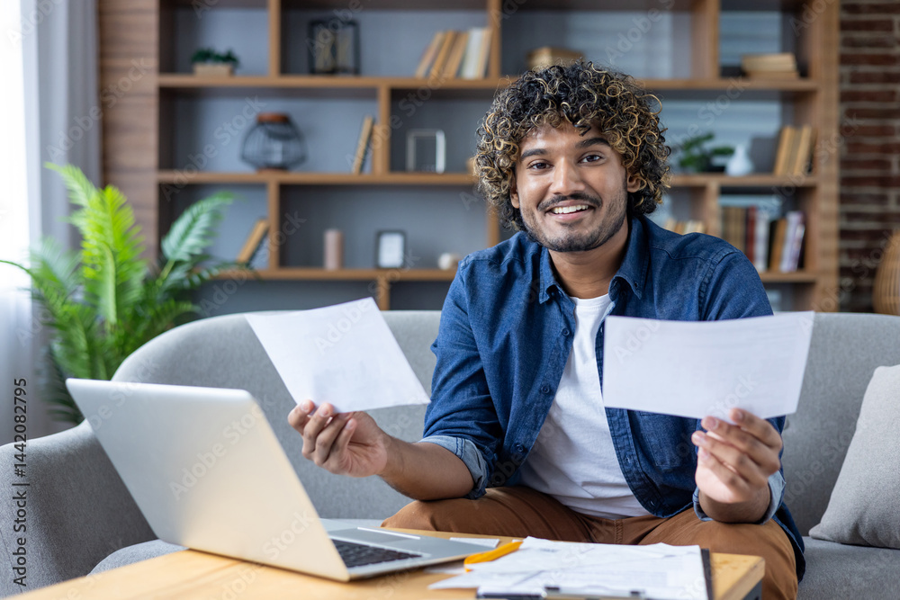A smiling man is looking at the camera holding papers in front of a laptop while working remotely from home.