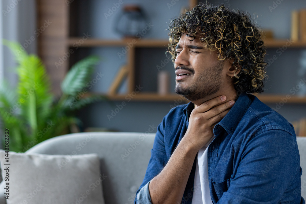 A man with curly hair holds his hand to his throat, seemingly in pain while sitting in a living room.