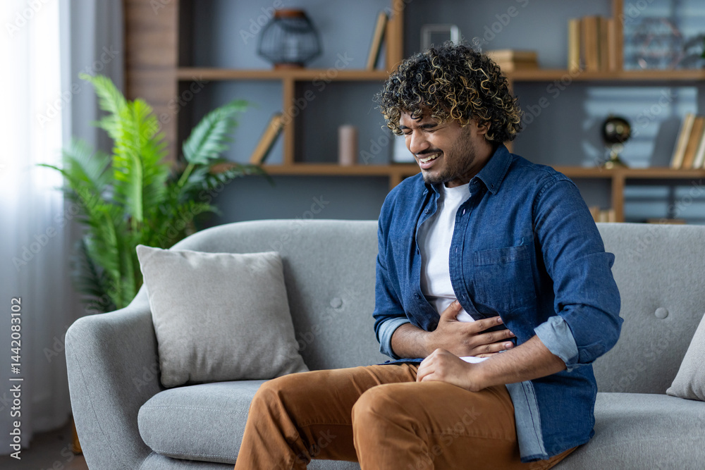 A young man sits on a sofa, experiencing stomach pain. He is holding his abdomen and appears to be in discomfort, possibly suffering from indigestion.