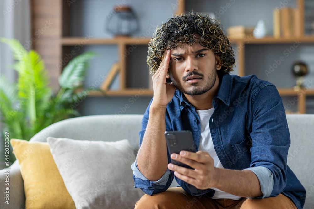 A concerned young man looks at his phone while sitting on a couch at home. He is holding his head with his hand.