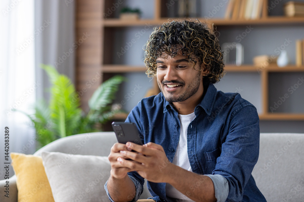 A young man with curly hair sits on a sofa, using a smartphone and smiling widely.