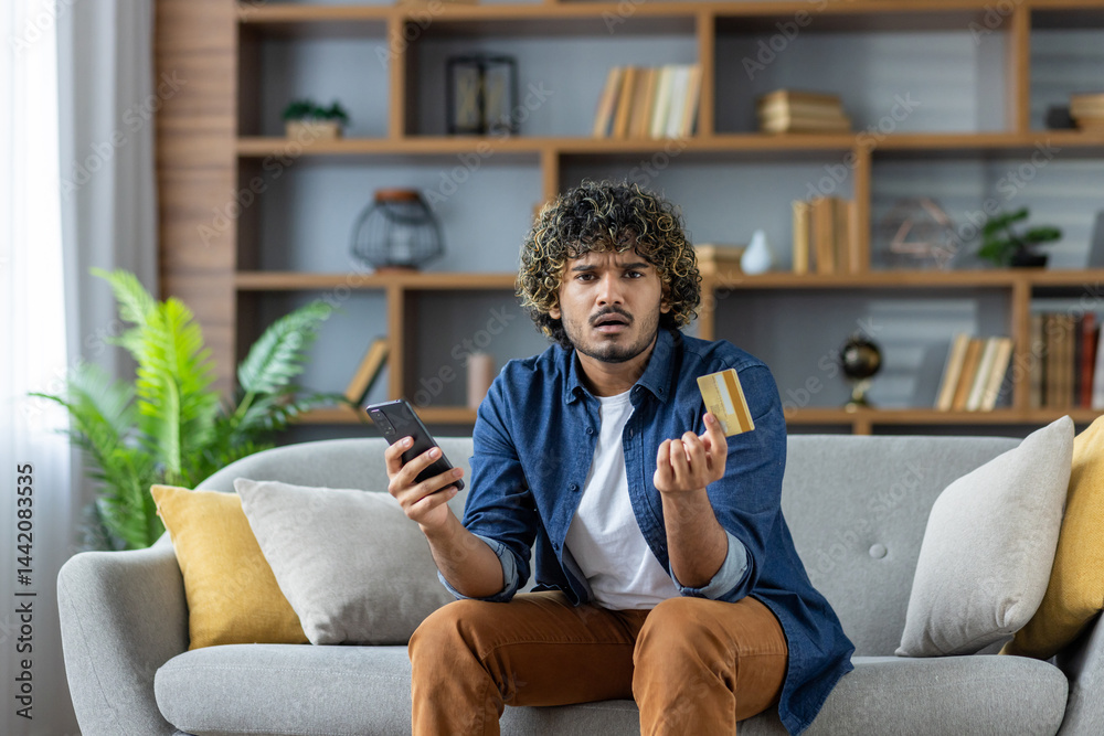 A concerned man sits on a couch, holding a credit card and a phone, likely experiencing payment issues. The room has a modern aesthetic.