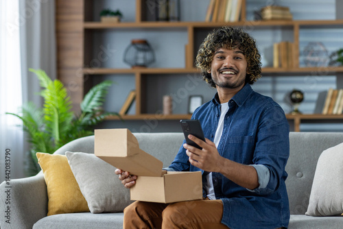 A smiling man holds a phone while opening a package on his couch. The background showcases a cozy living room environment.