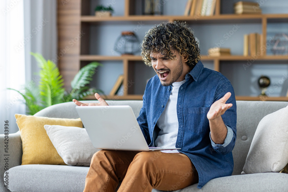 A frustrated man sits on a couch, looking at his laptop with a look of exasperation. The man wears casual clothes, indoors.