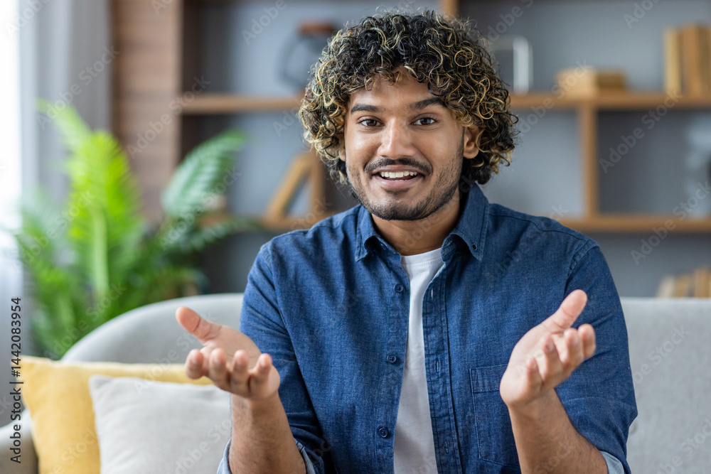 A smiling man with curly hair gestures with his hands while sitting on a sofa indoors. A comfortable living room setting.
