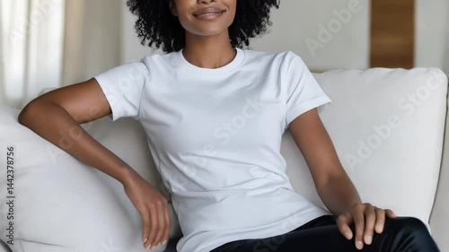 portrait of a young woman with curly hair, in a white t-chirt sitting on a coach,  A young woman exudes comfort and confidence as she sits in a cozy space filled with natural light. Her casual attire 
