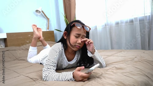 Asian girl looks tired on a cozy bed, holding a smartphone in a modern bedroom