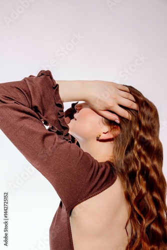 young woman with long wavy red hair touching her head with hand in stylish brown dress with open back against light background, beauty salon, haircare, fashion industry