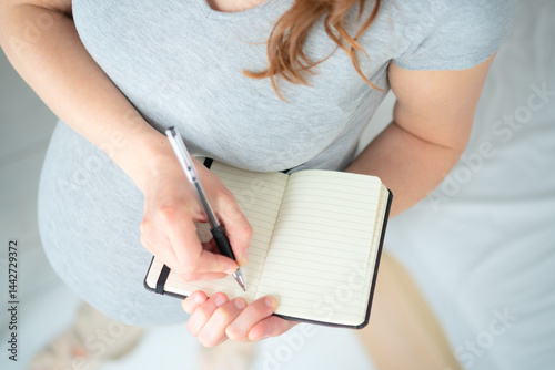 Future mother writing detailed maternity plan in notebook. Bird's-eye close-up shows careful preparation of hospital checklist and birth reminders.