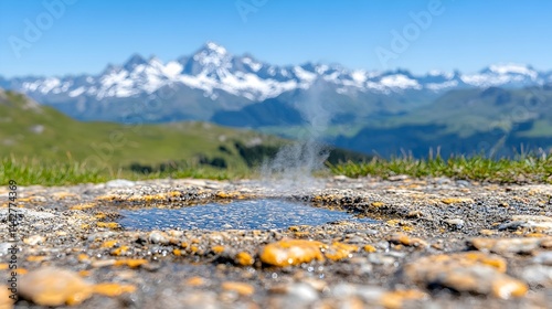 A small, steaming pool of water sits in a rocky area, with a majestic snow-capped mountain range in the background under a clear blue sky