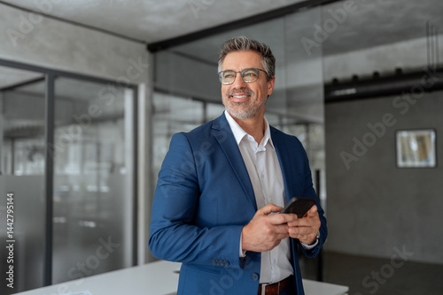 Smiling Indian mature businessman in suit holding smartphone standing in office, working on device. Executive professional ceo senior business manager man using cell phone mobile, looking dreaming